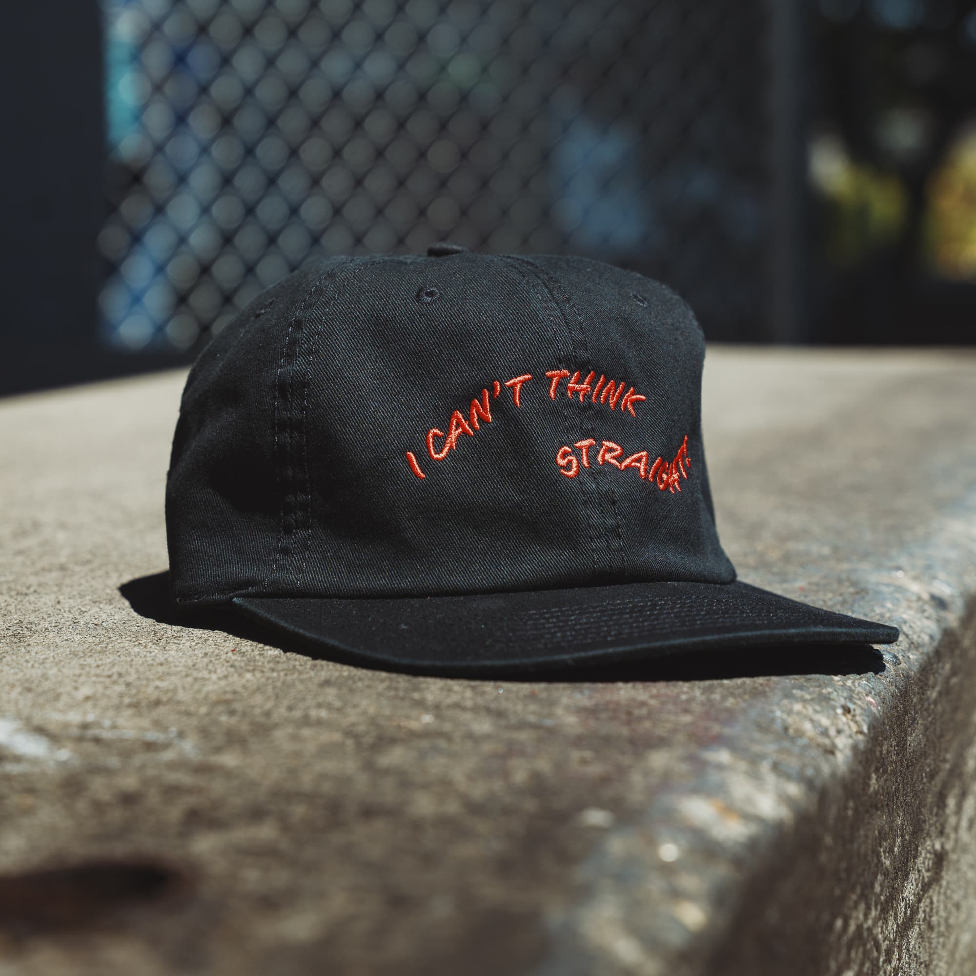 Black cap with red text 'I CAN'T THINK STRAIGHT' on a concrete surface with a blurred background.